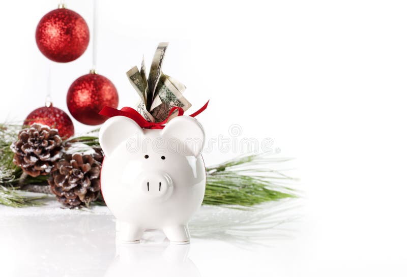 A white piggy bank tied with red ribbon and US currency in top slot, pig facing forward, christmas ornaments blurred in background. A concept for saving for the holidays as well as the expenses and costs of holidays. A white piggy bank tied with red ribbon and US currency in top slot, pig facing forward, christmas ornaments blurred in background. A concept for saving for the holidays as well as the expenses and costs of holidays.