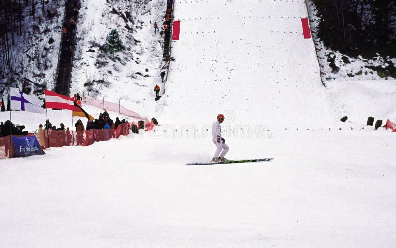 Chris broz stops at the bottom A numbered ski jumper just landed while others are climbing the steep stairs next to the tall hill and are waiting for their turn to compete at the top of 112 k kilometer hill at the annual westby wisconsin snowflake inetrnational ski jump tournament while the crowd of families at the bottom of the big snow covered hill in the united states of america. Chris broz stops at the bottom A numbered ski jumper just landed while others are climbing the steep stairs next to the tall hill and are waiting for their turn to compete at the top of 112 k kilometer hill at the annual westby wisconsin snowflake inetrnational ski jump tournament while the crowd of families at the bottom of the big snow covered hill in the united states of america