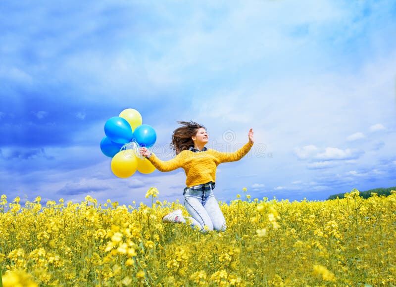 Jumping teenager girl in a field with airy blue and yellow balloons. A blue sky and a yellow field with blooming