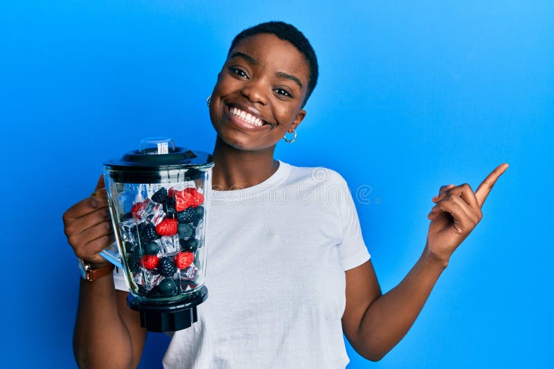Young african american woman holding food processor mixer machine with fruits smiling happy pointing with hand and finger to the side. Young african american woman holding food processor mixer machine with fruits smiling happy pointing with hand and finger to the side