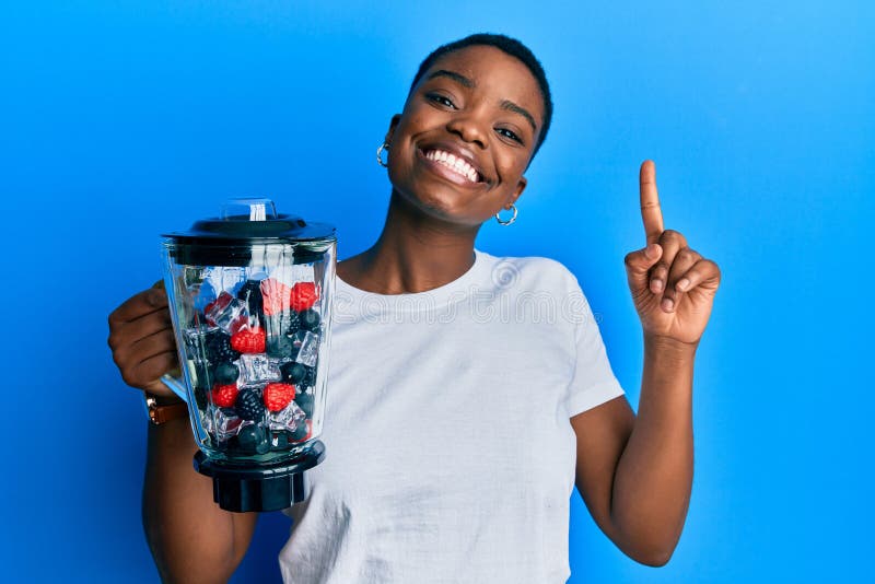 Young african american woman holding food processor mixer machine with fruits smiling with an idea or question pointing finger with happy face, number one. Young african american woman holding food processor mixer machine with fruits smiling with an idea or question pointing finger with happy face, number one