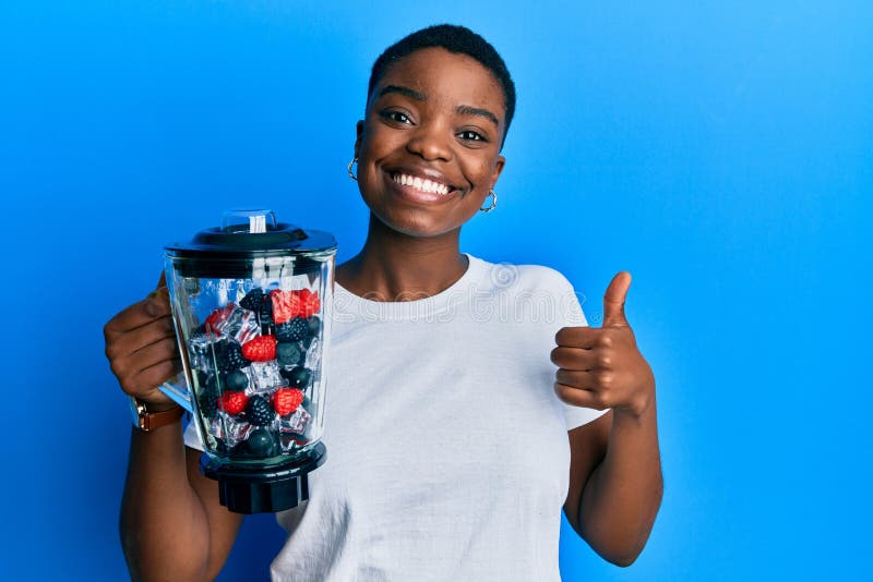 Young african american woman holding food processor mixer machine with fruits smiling happy and positive, thumb up doing excellent and approval sign. Young african american woman holding food processor mixer machine with fruits smiling happy and positive, thumb up doing excellent and approval sign