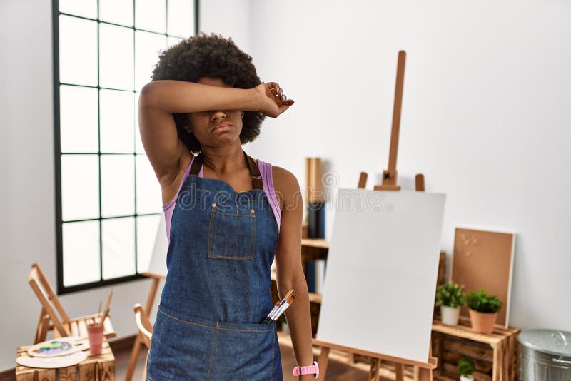 Young african american woman with afro hair at art studio covering eyes with arm, looking serious and sad. sightless, hiding and rejection concept. Young african american woman with afro hair at art studio covering eyes with arm, looking serious and sad. sightless, hiding and rejection concept