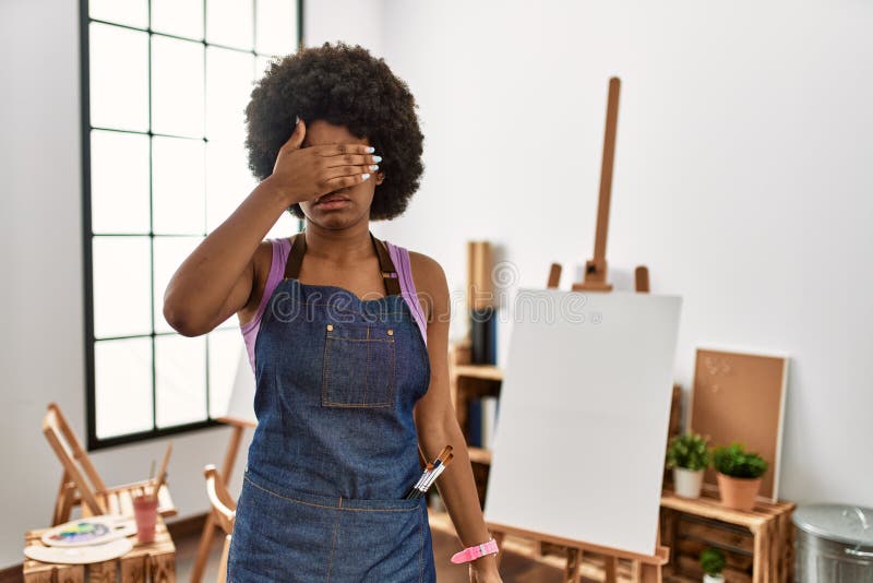 Young african american woman with afro hair at art studio covering eyes with hand, looking serious and sad. sightless, hiding and rejection concept. Young african american woman with afro hair at art studio covering eyes with hand, looking serious and sad. sightless, hiding and rejection concept