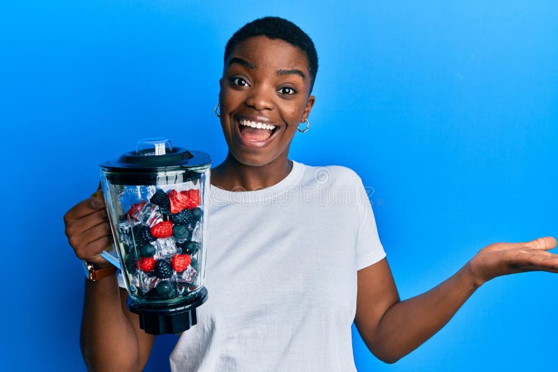 Young african american woman holding food processor mixer machine with fruits celebrating achievement with happy smile and winner expression with raised hand. Young african american woman holding food processor mixer machine with fruits celebrating achievement with happy smile and winner expression with raised hand
