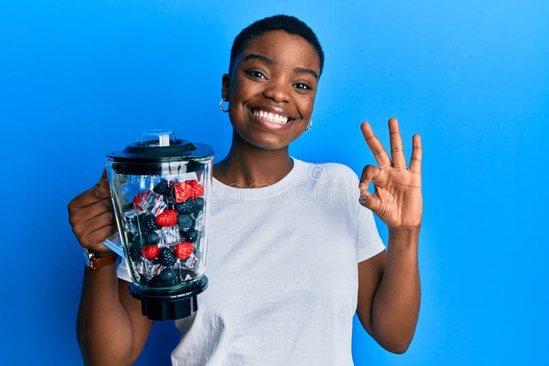 Young african american woman holding food processor mixer machine with fruits doing ok sign with fingers, smiling friendly gesturing excellent symbol. Young african american woman holding food processor mixer machine with fruits doing ok sign with fingers, smiling friendly gesturing excellent symbol