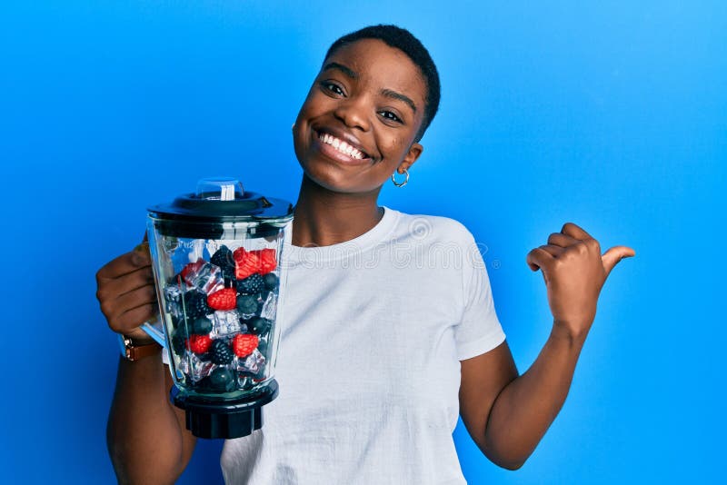 Young african american woman holding food processor mixer machine with fruits pointing thumb up to the side smiling happy with open mouth. Young african american woman holding food processor mixer machine with fruits pointing thumb up to the side smiling happy with open mouth