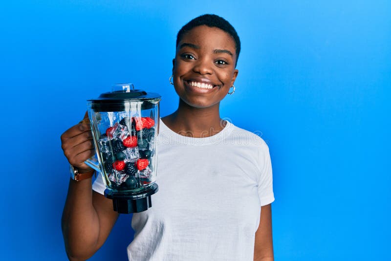 Young african american woman holding food processor mixer machine with fruits looking positive and happy standing and smiling with a confident smile showing teeth. Young african american woman holding food processor mixer machine with fruits looking positive and happy standing and smiling with a confident smile showing teeth