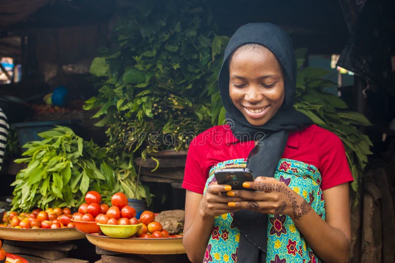 Junge afrikanische Frau, die auf einem lokalen Markt verkauft, lächelt, wenn sie ihr Mobiltelefon benutzt