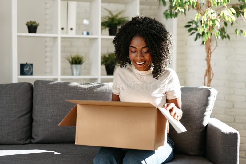 Junge afrikanische Frau sitzt zu Hause auf einem Sofa und packt Pappkarton mit Online-Einkauf aus stockbilder