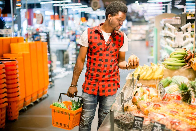 Young african man buying vegetables and fruits in grocery section at supermarket. Black man choose vegetables and fruits in the supermarket while holding grocery basket. Man shopping veggies at supermarket. Young african man buying vegetables and fruits in grocery section at supermarket. Black man choose vegetables and fruits in the supermarket while holding grocery basket. Man shopping veggies at supermarket