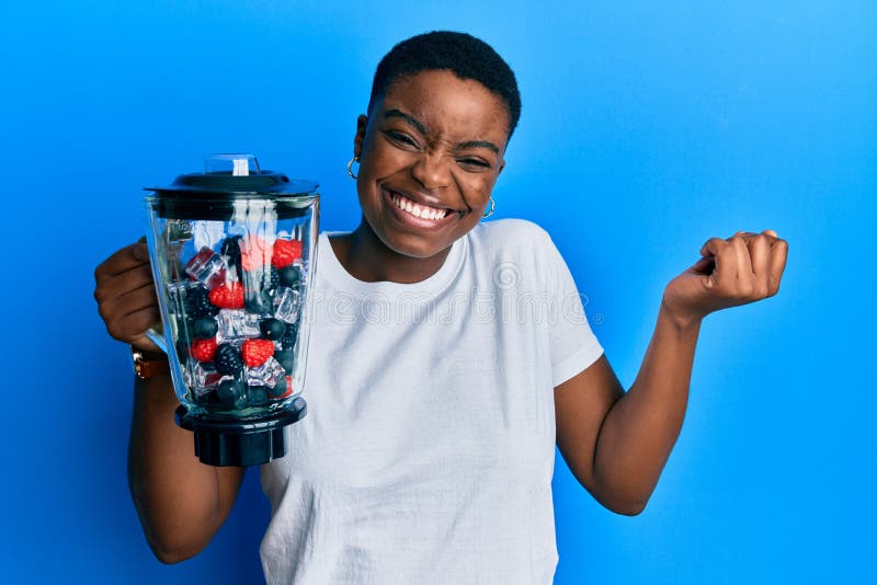 Young african american woman holding food processor mixer machine with fruits screaming proud, celebrating victory and success very excited with raised arm. Young african american woman holding food processor mixer machine with fruits screaming proud, celebrating victory and success very excited with raised arm