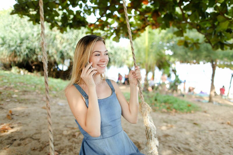 Junge Amerikanerin Reiten Schaukel und sprechen mit Smartphone Sand und Baum im Hintergrund.