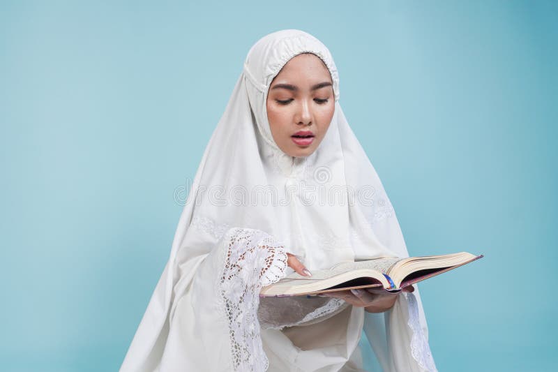 Junge asiatische Muslimfrau im Gebetskleid sitzt herunter und liest Quran über isolierten blauen Hintergrund. Hadsch-Konzept stockfoto