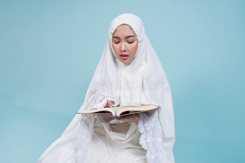 Junge asiatische Muslimfrau im Gebetskleid sitzt herunter und liest Quran über isolierten blauen Hintergrund. Hadsch-Konzept stockfotografie