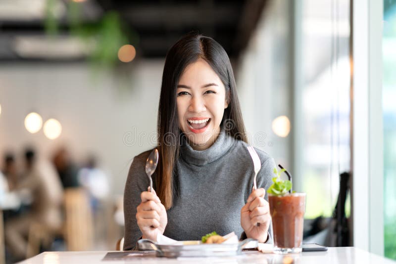 Young attractive asian woman holding fork and spoon feeling hungry, excited, happy and ready to eat healthy food looking at camera in cafe coffee shop. Charming blogger or social influencer concept. Young attractive asian woman holding fork and spoon feeling hungry, excited, happy and ready to eat healthy food looking at camera in cafe coffee shop. Charming blogger or social influencer concept.