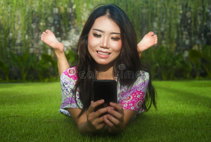 Junge attraktive schöne und herrliche asiatische Chinesin, die auf Park des grünen Grases unter Verwendung Internet-Social Media- lizenzfreie stockfotos Junge attraktive schöne und herrliche asiatische Chinesin, die auf Park des grünen Grases unter Verwendung Internet-Social Media- lizenzfreie stockfotos