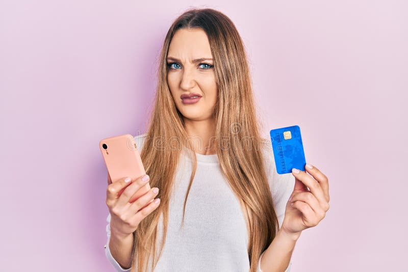 Young blonde girl holding smartphone and credit card clueless and confused expression. doubt concept. Young blonde girl holding smartphone and credit card clueless and confused expression. doubt concept
