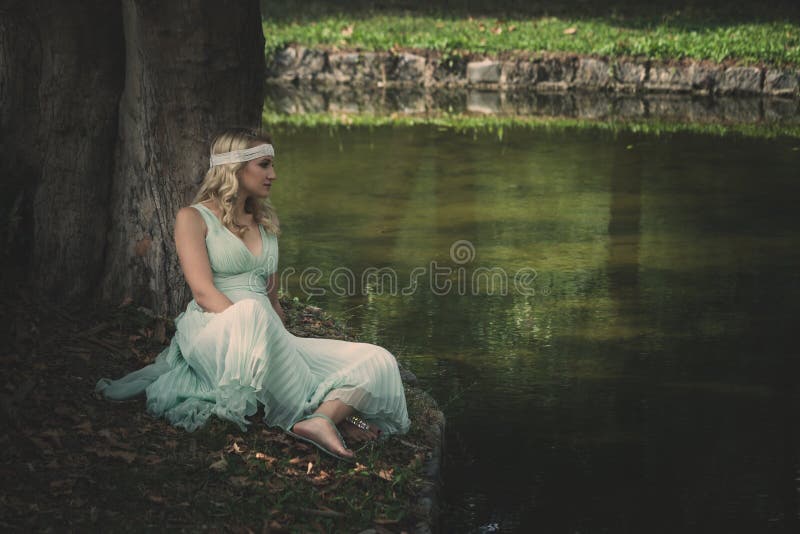 Young blonde woman in romantic dress and hair decoration sit by lake lean on tree summer day. Young blonde woman in romantic dress and hair decoration sit by lake lean on tree summer day