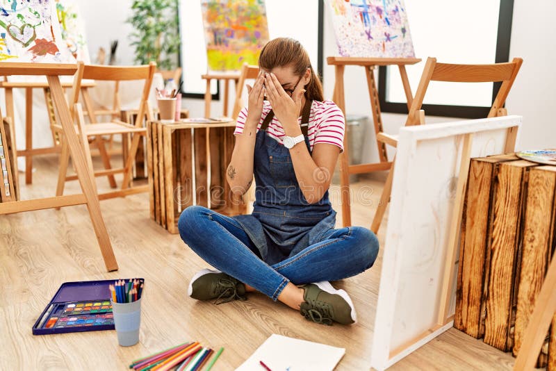 Young brunette woman at art studio sitting on the floor with sad expression covering face with hands while crying. depression concept. Young brunette woman at art studio sitting on the floor with sad expression covering face with hands while crying. depression concept