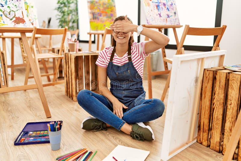 Young brunette woman at art studio sitting on the floor smiling and laughing with hand on face covering eyes for surprise. blind concept. Young brunette woman at art studio sitting on the floor smiling and laughing with hand on face covering eyes for surprise. blind concept