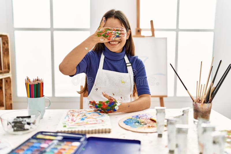 Young brunette woman at art studio with painted hands smiling and laughing with hand on face covering eyes for surprise. blind concept. Young brunette woman at art studio with painted hands smiling and laughing with hand on face covering eyes for surprise. blind concept