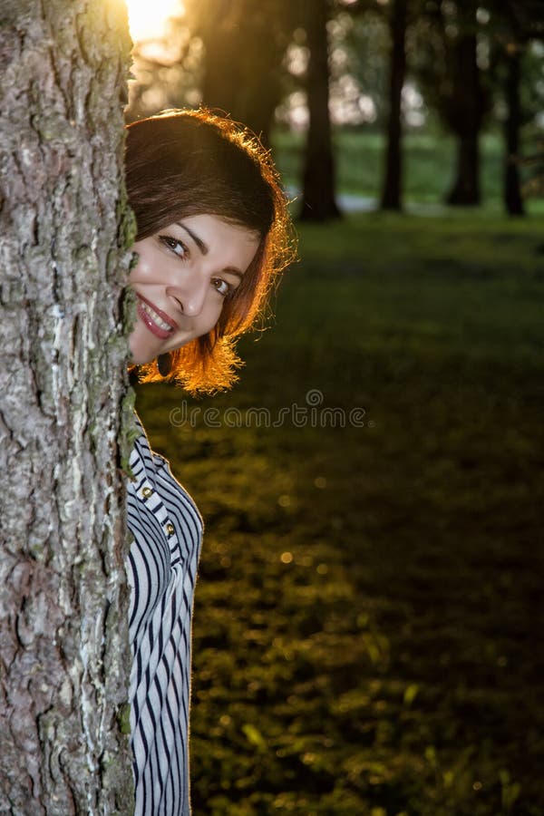 Junge Brunettefrau knallt heraus hinter dem Baum durch Sonnenuntergang lizenzfreie stockfotografie Junge Brunettefrau knallt heraus hinter dem Baum durch Sonnenuntergang lizenzfreie stockfotografie