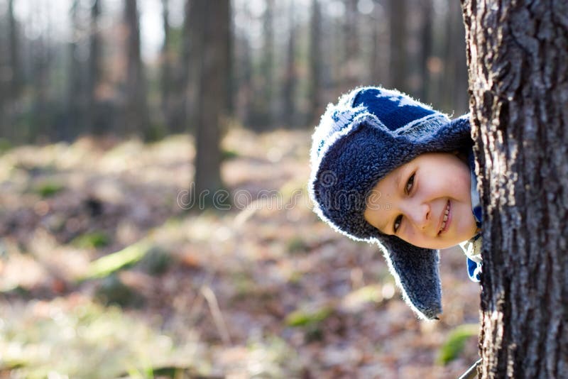 A smiling boy, wearing a blue winter hat, peeks his head around a tree while hiding and playing in a forest on a cold day. A smiling boy, wearing a blue winter hat, peeks his head around a tree while hiding and playing in a forest on a cold day.