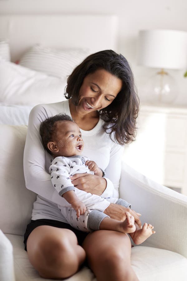 Junge erwachsene Mutter, die in einem Lehnsessel in ihrem Schlafzimmer sitzt, ihren dreimonatigen alten Babysohn in ihren Armen h stockfotografie Junge erwachsene Mutter, die in einem Lehnsessel in ihrem Schlafzimmer sitzt, ihren dreimonatigen alten Babysohn in ihren Armen h stockfotografie