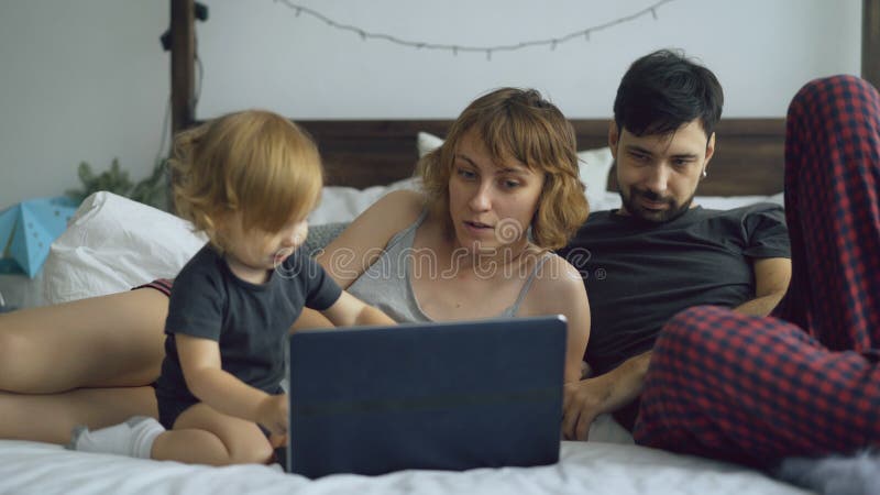 Junge Familie mit dem netten kleinen Mädchen, das mit Tochter während sie schreibend auf der Laptop-Computer zu Hause sitzt im Be lizenzfreies stockfoto