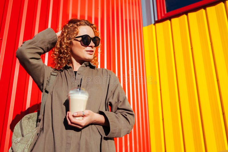 Junge Frau des Hippies mit trinkendem Kaffee des gelockten roten Haares gegen rote und gelbe Wand Tourist mit dem Rucksackkühlen lizenzfreies stockfoto Junge Frau des Hippies mit trinkendem Kaffee des gelockten roten Haares gegen rote und gelbe Wand Tourist mit dem Rucksackkühlen lizenzfreies stockfoto
