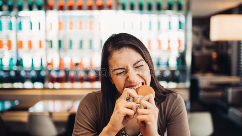 Junge Frau, die fetthaltigen Hamburger isst Sehnender Schnellimbiß Das schuldige Vergnügen, ungesunde Fertigkost essend genießen