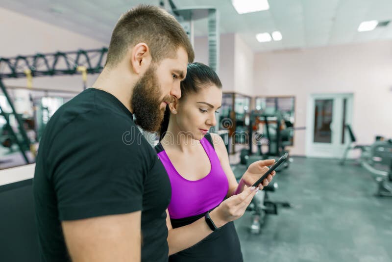 Junge Frau, die mit persönlichem Trainer in der Turnhalle, schauend im Smartphone und in der Diskussion spricht Eignung, Sport, T lizenzfreies stockbild Junge Frau, die mit persönlichem Trainer in der Turnhalle, schauend im Smartphone und in der Diskussion spricht Eignung, Sport, T lizenzfreies stockbild