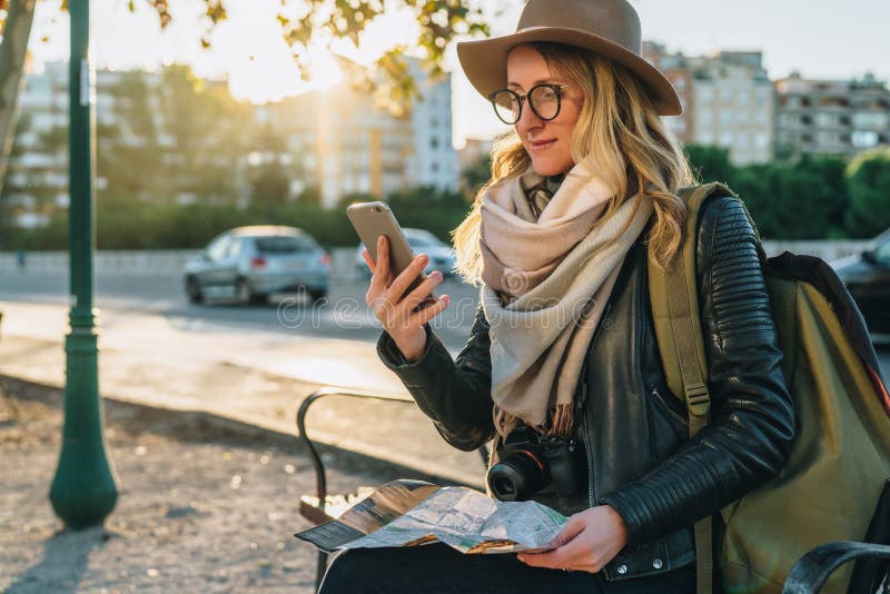 Junge Frau, Hippie-Mädchentourist mit Rucksack und Kamera sitzt auf Bank in der Stadtstraße, benutzt den Smartphone und hält Kart lizenzfreies stockfoto Junge Frau, Hippie-Mädchentourist mit Rucksack und Kamera sitzt auf Bank in der Stadtstraße, benutzt den Smartphone und hält Kart lizenzfreies stockfoto