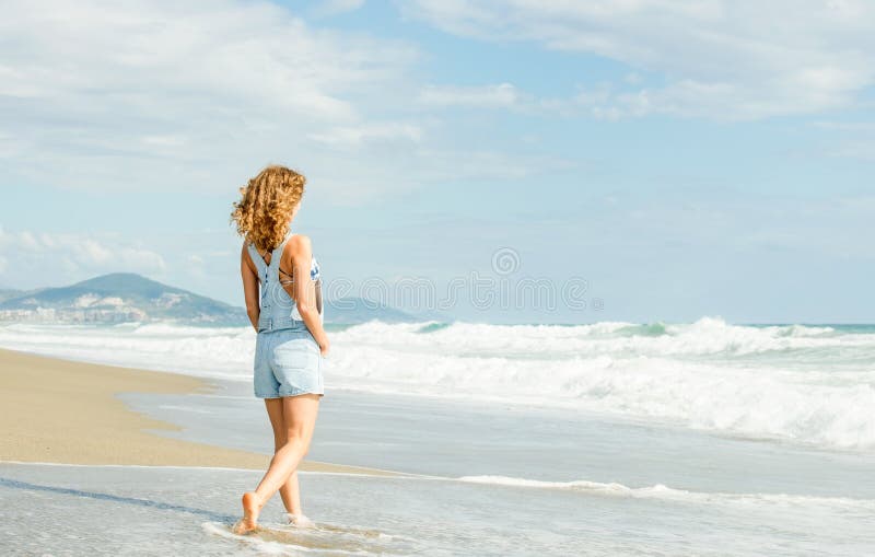 Junge Frau im blauen Denimoverall gehend entlang Strand und der stürmische Ozean am sonnigen Tag stockbilder Junge Frau im blauen Denimoverall gehend entlang Strand und der stürmische Ozean am sonnigen Tag stockbilder