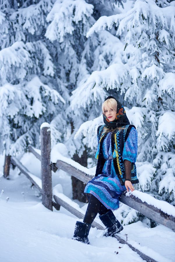 Full length portrait of fashionable woman in blue dress, embroidered warm sleeveless fur coat and kerchief with floral design outdoor at wooden fence on snowy spruce tree brunches blurred background. Full length portrait of fashionable woman in blue dress, embroidered warm sleeveless fur coat and kerchief with floral design outdoor at wooden fence on snowy spruce tree brunches blurred background.