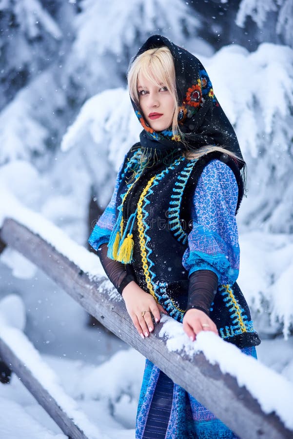 Young attractive fashionable blond woman in blue dress, embroidered warm sleeveless fur coat and kerchief with floral design outdoor at wooden fence on snowy spruce tree brunches blurred background. Young attractive fashionable blond woman in blue dress, embroidered warm sleeveless fur coat and kerchief with floral design outdoor at wooden fence on snowy spruce tree brunches blurred background.