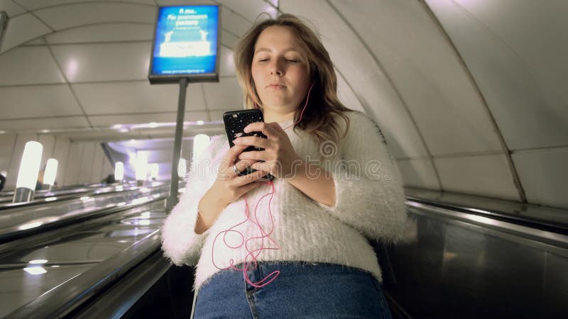 Junge Frau in Kopfhörer auf Rolltreppe in der Metro. Medien. Unteransicht einer schönen Frau, die in die U-Bahn absteigt. Frau