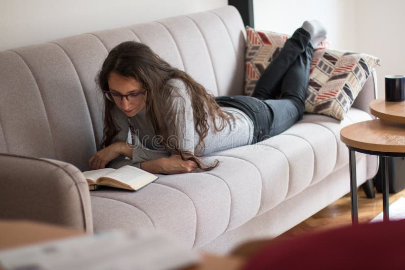 Junge Frau liegt auf dem Sofa und liest ein interessantes Buch lizenzfreie stockfotografie