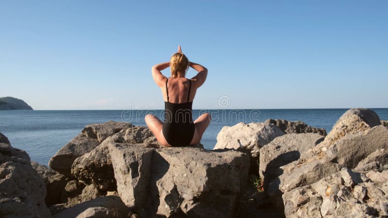 Junge Frau meditiert in einem schwarzen Badeanzug. Frau praktiziert Yoga mit Blick auf das Meer oder das Meer bei Sonnenuntergang stock video footage
