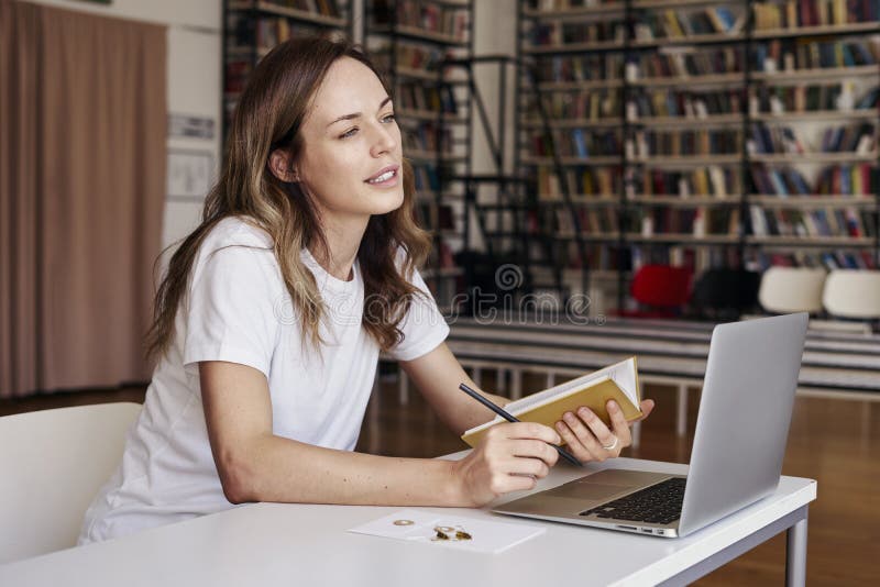 Junge Frau mit dem langen Haar, das hinten an Laptop an mit-arbeitendem Büro oder an Bibliothek, Bücherregal arbeitet Geschäftsfr stockfotografie