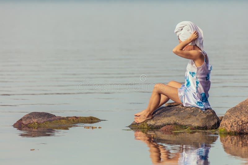 Junge Frau mit einem Handtuch auf dem Kopf am Meer. Frau sitzt auf einem Stein auf dem Hintergrund des Meeres. Rücken. Spa-Konzept stockfotografie