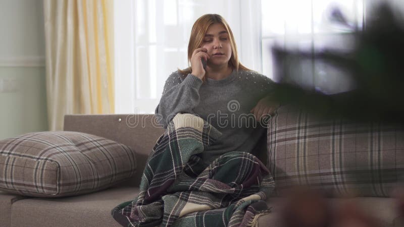 Junge Frau Plys-Größe, die beim Sprechen am intelligenten Telefon zuhause sitzt onder Decke auf dem Sofa lächelt Plusgrößenfrau
