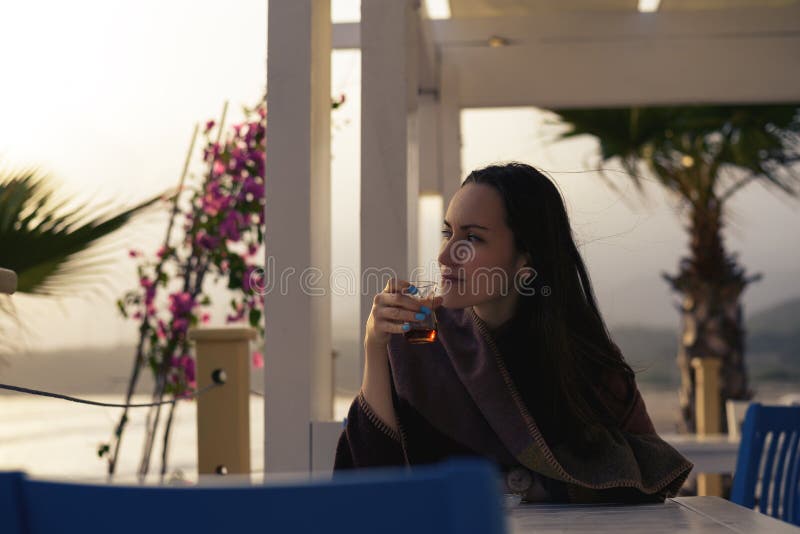 Junge Frau sitzt an einem Tisch auf einer Sommerterrasse des Cafés auf der Küste trinkt Tee in einem Plaid lizenzfreies stockfoto Junge Frau sitzt an einem Tisch auf einer Sommerterrasse des Cafés auf der Küste trinkt Tee in einem Plaid lizenzfreies stockfoto