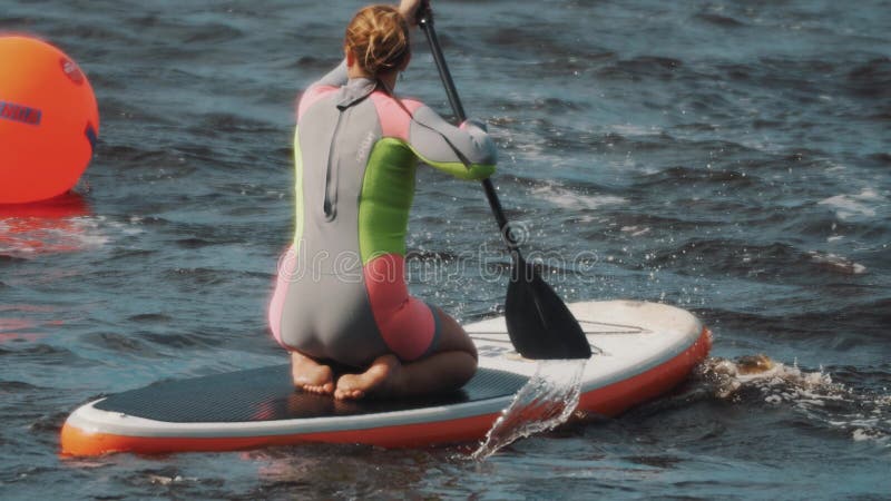 Junge Frauen im bunten Badeanzug sitzt auf einem Surfbrett und reitet es unter Verwendung des Paddels