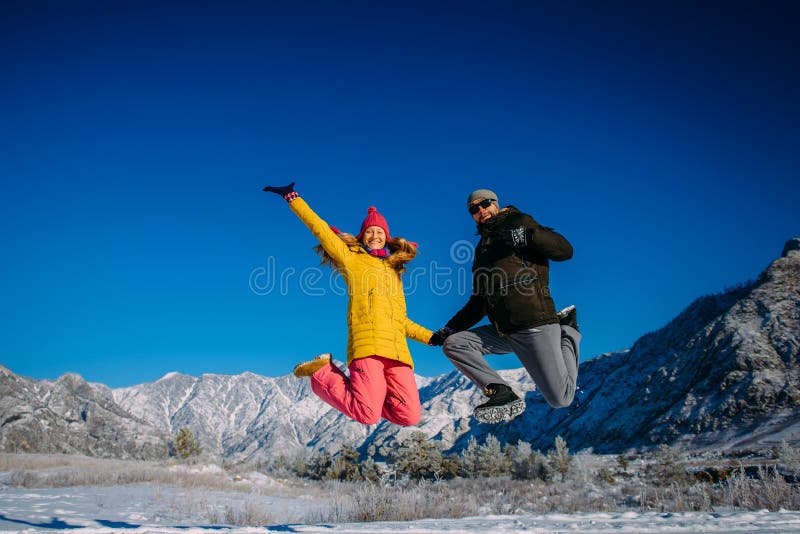 Junge fröhliche Paare in hellen Kleider springen am sonnigen Wintertag auf den Hintergrund schneebedeckter Berge Junge und Mädche lizenzfreies stockfoto Junge fröhliche Paare in hellen Kleider springen am sonnigen Wintertag auf den Hintergrund schneebedeckter Berge Junge und Mädche lizenzfreies stockfoto