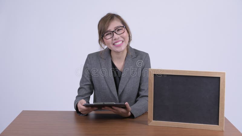 Junge glückliche asiatische Geschäftsfrau, die etwas mit Tafel am Schreibtisch zeigt