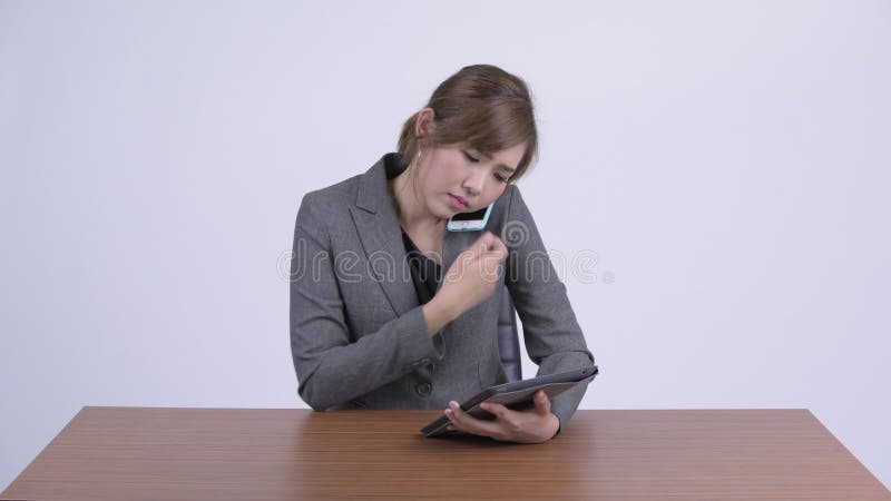 Junge glückliche asiatische Geschäftsfrau, die am Telefon bei der Anwendung der digitalen Tablette bei der Arbeit spricht