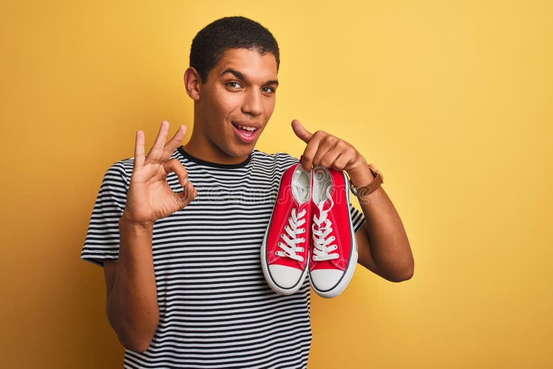 Junge, gut aussehende Arab-Mann, der lockere Sneakers über isoliertem gelbem Hintergrund hält und mit den Fingern hervorragend sig stockbilder