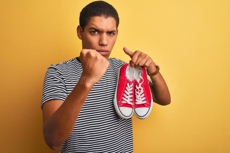 Junge, gut aussehende Arab-Mann, der lockere Sneakers über isoliertem gelbem Hintergrund hält, ärgerlich und frustriert mit einem stockfotografie